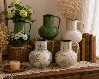 Pode incluir: Cinco jarros de cerâmica com pegas, apresentando designs variados incluindo verde sólido, branco com flores azuis, branco com folhagem verde e verde com margaridas brancas. Os jarros têm um acabamento texturizado e bordos escuros.