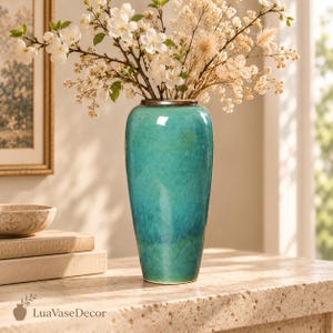 Könnte beinhalten: Eine hohe, zylindrische Keramikvase mit einer glatten, glänzenden türkisen und blaugrünen Farbverlaufglasur. Die Vase hat einen dunklen, schmalen Rand und ist mit einem Strauß weißer Kirschblüten und getrockneten beigen Gräsern gefüllt, inklusive Text LuaVaseDecor.