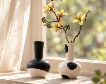 Pode incluir: Dois vasos de cerâmica com acabamento fosco. Um vaso é preto sólido na metade superior e branco na inferior. O outro é branco com manchas circulares pretas. Um ramo com flores amarelas e botões verdes está no vaso manchado. O texto LuaVaseDecor é visível.