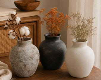 May include: Three rounded vases with textured finishes in light gray, dark charcoal, and off-white. The gray vase holds cotton stems, the charcoal vase contains orange dried flowers, and the white vase holds light beige dried sprigs.