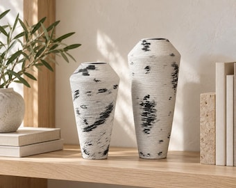 Pode incluir: Dois vasos cónicos com uma superfície branca texturizada e padrões irregulares de pinceladas pretas horizontais. Os vasos têm um pescoço estreito e um corpo mais largo, colocados lado a lado sobre uma superfície plana.