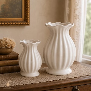 May include: Two white ceramic vases with a vertical ribbed texture and a ruffled, scalloped rim. The vases feature a bulbous body that tapers towards the neck and flares at the top. The set includes one smaller vase and one larger vase.