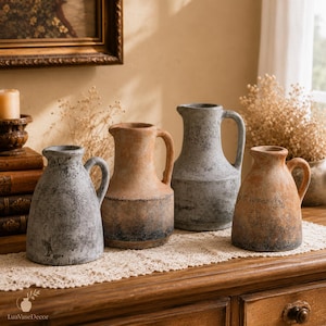 May include: Four ceramic pitchers with textured, weathered finishes in shades of gray and terracotta. Each pitcher features a rounded body, a short spout, and a single side handle. The surfaces display a mottled, rustic appearance with dark accents.