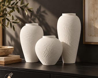 Pode incluir: Três vasos em tom branco-sujo com acabamento de superfície texturizado e rugoso. O conjunto inclui dois vasos altos e esguios e um vaso mais curto e arredondado, todos com gargalo estreito e abertura circular no topo.