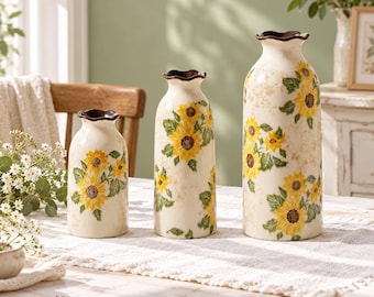 Pode incluir: Um conjunto de três vasos de cerâmica na cor creme com alturas variadas, cada um apresentando um padrão de girassóis amarelos com folhas verdes e uma borda recortada em marrom escuro. Os vasos estão alinhados sobre um caminho de mesa.