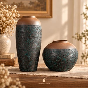 May include: Two ceramic vases with a smooth tan upper section and a textured, dark brown and teal mottled lower body. The taller vase is cylindrical, while the shorter vase is rounded. The text LuaVaseDecor is printed at the bottom centre.