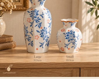 Pode incluir: Dois vasos de cerâmica branca com padrões florais azuis e laranja e bordas recortadas. O vaso grande tem 24 cm de altura e o pequeno 15 cm. Um guia de tamanhos lista as dimensões de altura, abertura e base para ambos os vasos.