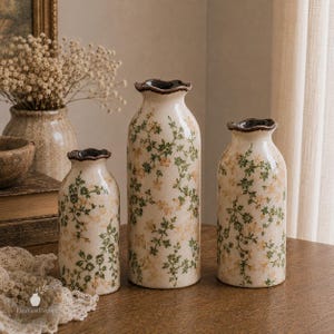 May include: Three ceramic vases with a cream-coloured base, featuring a delicate pattern of small green leaves and light yellow flowers. Each vase has a cylindrical body, a narrow neck, and a dark brown, scalloped, ruffled rim edge.