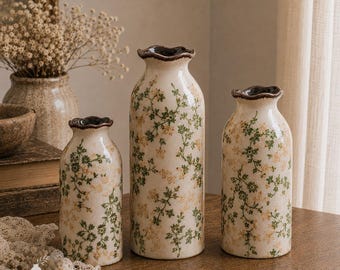 Pode incluir: Três vasos de cerâmica com base cor de creme, apresentando um padrão delicado de pequenas folhas verdes e flores amarelo-claras. Cada vaso tem um corpo cilíndrico, um pescoço estreito e uma borda superior ondulada em castanho escuro.