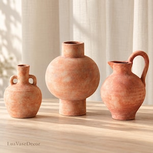 May include: Three terracotta-colored ceramic vessels with a textured, matte finish. The set includes a small vase with two side handles, a large spherical vase with a wide neck, and a pitcher featuring a curved handle and a spout.