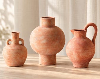 Pode incluir: Três recipientes de cerâmica na cor terracota com acabamento fosco e texturizado. O conjunto inclui um vaso pequeno com duas alças laterais, um vaso esférico grande de pescoço largo e uma jarra com alça curva e bico.