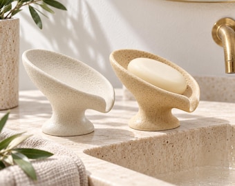 Pode incluir: Dois saboneteiras de cerâmica salpicada com um design curvo e elevado. Uma saboneteira é de cor branca suja e está vazia, enquanto a outra é de cor castanho claro e contém um sabonete redondo, liso e de cor creme. Ambas têm acabamento texturizado.