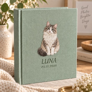 May include: A sage green fabric-covered photo album featuring an embroidered illustration of a brown and white tabby cat. Below the cat, the name LUNA and the date 03.12.2024 are embroidered in dark green thread on the front cover of the album.