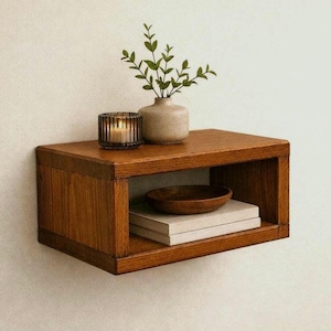 Puede incluir: Un estante de pared rectangular de madera con acabado marrón medio. Encima hay un pequeño jarrón de cerámica beige con hojas verdes y un portavelas de cristal estriado. Dentro del estante, un cuenco de madera oscura descansa sobre dos libros blancos.