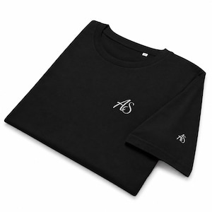 Könnte beinhalten: Ein schwarzes Kurzarm-T-Shirt mit einem weißen, gestickten AS-Schriftzug auf der linken Brustseite und einem kleineren, passenden AS-Logo am Rand des rechten Ärmels. Das T-Shirt hat einen Rundhalsausschnitt und ein eingenähtes Größenetikett.
