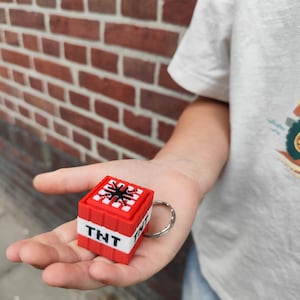Op de afbeelding: Een rood-witte kubusvormige sleutelhanger met de tekst TNT in het zwart op een witte band. De bovenkant van de kubus toont een zwarte ster-afbeelding op een wit rasterpatroon. Een zilveren metalen ring is aan de zijkant van het blok bevestigd.