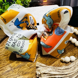 Puede incluir: Un lazo grande para el pelo hecho de cinta de grosgrain blanca con un estampado gráfico repetitivo de un personaje de paloma azul, detalles en naranja y el texto "The Pigeon HAS to Go to School!" de Mo Willems, atado con un nudo central.