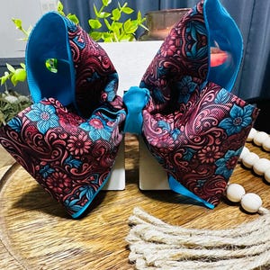 Puede incluir: Un lazo grande para el cabello hecho de cinta de grosgrain con un patrón vibrante de flores azul cerceta y diseños de cachemira en magenta oscuro. El lazo está centrado con un nudo de cinta azul brillante y tiene bordes azul brillante.