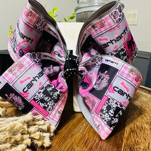 Op de afbeelding: Een grote haarstrik van grosgrain lint met een herhalend patroon in roze, zwart en wit. De print bevat de tekst Howdy Carhartt, Nashville en koeienprintmotieven. Een klein middenstuk van zwart-wit gestippeld lint.