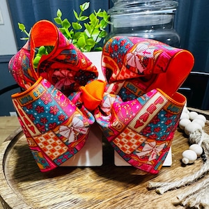 May include: A large hair bow made of vibrant grosgrain ribbon featuring a patchwork pattern of bright pink, teal, and orange squares. The design includes motifs of white bows, small flowers, and the word LOVE in block letters, tied with an orange centre.