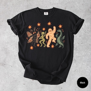 May include: A black short-sleeve t-shirt featuring a graphic print of a moth, an alien, a yeti, and a sea monster dancing among orange stars. The design is centered on the chest, and the shirt has a crew neckline and rolled sleeves.