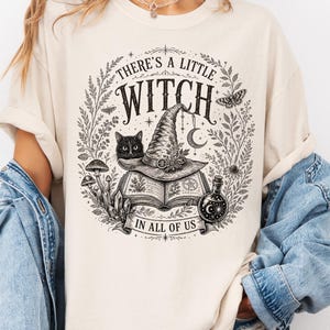 May include: An ivory t-shirt featuring a black graphic print of a witch hat resting on an open book, a black cat, mushrooms, crystals, a potion bottle, and the text "THERE'S A LITTLE WITCH IN ALL OF US" surrounded by a circular botanical wreath.