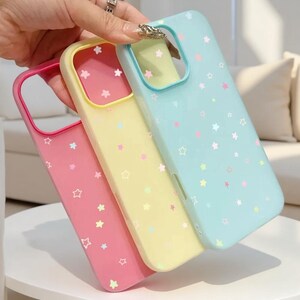 May include: Three smartphone cases in pink, yellow, and light blue, each featuring a pattern of small, scattered stars in various pastel colors. The cases have a rectangular cutout for a camera module and a smooth, matte-finish surface.