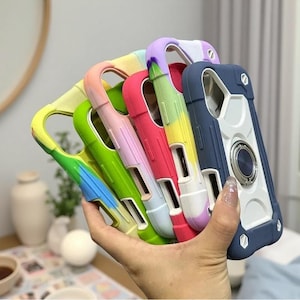 May include: A collection of five protective smartphone cases held in a hand. The cases feature a rugged design with reinforced corners, cutouts for camera lenses, and a circular metal ring stand on the back of the dark blue case.