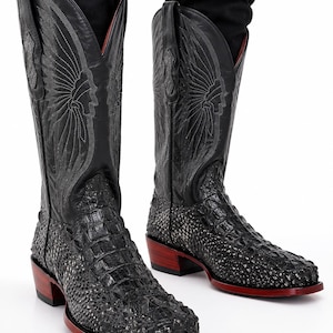 Puede incluir: Un par de botas vaqueras de cuero negro con un empeine texturizado de patrón reptiliano y una caña alta con detalladas costuras que forman una silueta de perfil. Las botas tienen un tacón de madera con una base de tono rojizo.