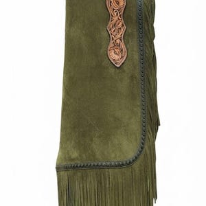 May include: Olive green suede chaps featuring tan leather panels with intricate floral tooling at the top and centre. The garment includes a dark leather border with decorative stitching and long, flowing fringe along the outer side edge.
