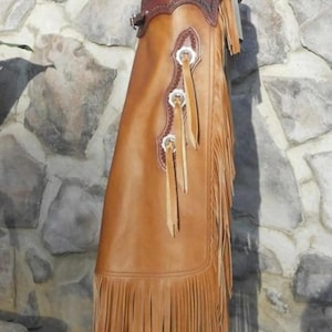 May include: A tan leather chap featuring a dark brown leather yoke at the top with decorative stitching. The side panel includes three silver-toned conchos with leather fringe accents, and the bottom edge is finished with long, vertical leather fringe.