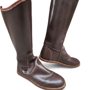 May include: A pair of knee-high dark brown boots featuring a smooth leather-like finish, rounded toes, and a flat sole. The boots are accented with light tan contrast stitching along the seams and a matching tan trim around the top opening.