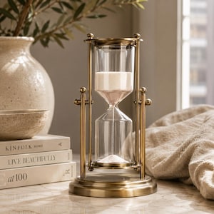 Puede incluir: Un reloj de arena giratorio con un marco de metal de latón cepillado y un recipiente de vidrio transparente lleno de arena blanca fina. El reloj de arena está montado sobre una base circular con pilares de soporte y un mecanismo de pivote.