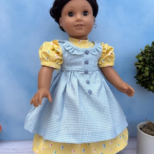 May include: A doll with dark brown hair styled in a braided crown, wearing a yellow short-sleeved dress with a colorful dot pattern and a light blue gingham pinafore featuring a ruffled neckline and a vertical row of four small blue buttons.