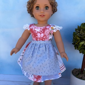 May include: A doll with wavy brown hair and brown eyes wearing a dress with a red floral bodice, white lace cap sleeves, and a light blue apron featuring a small floral pattern over a multi-patterned skirt with red, white, and blue fabric panels.