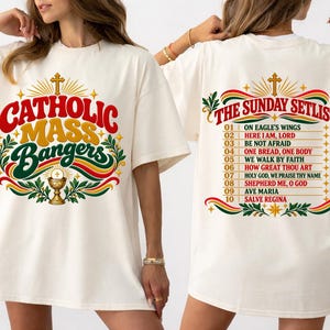 May include: An ivory t-shirt featuring a graphic print. The front displays the text CATHOLIC MASS BANGERS with a chalice and cross. The back lists THE SUNDAY SETLIST with ten song titles including On Eagle's Wings and Salve Regina.
