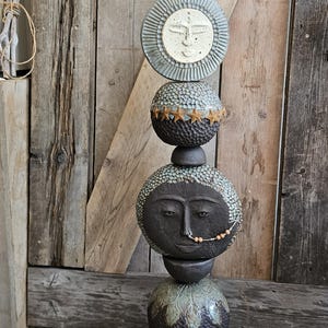 May include: A vertical ceramic totem sculpture composed of stacked, textured spheres and discs. The piece features a central face with a beaded nose ring, a sun-like disc with a face, and a top floral element, all finished in dark, earthy tones.