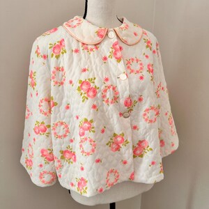 May include: A white quilted jacket featuring a repeating pattern of pink roses and green leaves. The jacket has a rounded collar with pink trim, three-quarter length sleeves, and a front button closure with three white buttons on a mannequin.