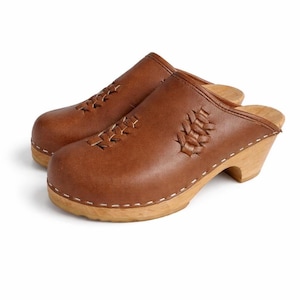 May include: A pair of brown leather clogs featuring a rounded toe, a low wooden heel, and decorative woven leather detailing across the top. The upper is attached to the light-colored wooden sole with visible white stitching along the base.
