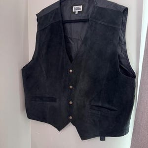 May include: A black suede vest featuring a V-neckline, four front buttons, and two lower welt pockets. The interior label reads Basic Editions. The vest is sleeveless with a smooth lining and is displayed on a black plastic hanger.