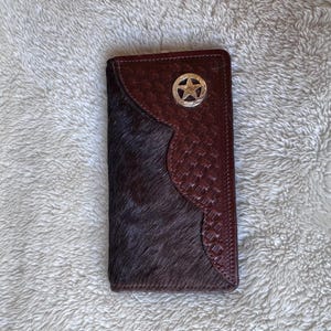 May include: A rectangular wallet featuring a dark brown hair-on-hide panel on the left side and a dark brown embossed leather panel on the right side. A silver-toned circular metal emblem with a five-pointed star is centered on the leather.