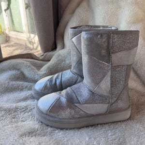 May include: A pair of mid-calf boots featuring a patchwork design of silver metallic and glittery panels. The boots have a rounded toe, a thick white rubber sole, and a soft, light-colored interior lining visible at the top opening.