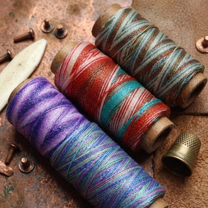 May include: Three spools of variegated sewing thread featuring distinct colour palettes: one purple and blue, one red and teal, and one teal and copper. The spools are arranged alongside small metal rivets, a bone folder, and a brass thimble.