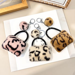May include: Three fuzzy keychains shaped like miniature handbags, each featuring a leopard print pattern in pink, white, or brown. Each keychain includes a matching plush pom-pom, a black circular handle, and a silver-toned metal lobster clasp.