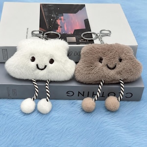 May include: Two plush cloud keychains, one white and one brown, each featuring embroidered dark eyes and a smiling mouth. Both keychains have two dangling black and white twisted cords ending in small fuzzy pom-poms and a silver metal ring.