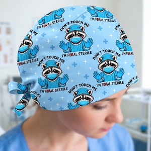 May include: A light blue scrub cap featuring a repeating pattern of a cartoon raccoon wearing a surgical cap and mask with raised hands. The text reads DON'T TOUCH ME I'M FERAL STERILE, with the word FERAL having a red line through it.
