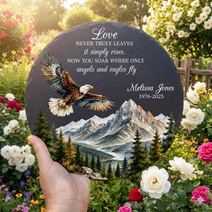 May include: A round slate memorial plaque featuring a printed illustration of a bald eagle in flight over a mountain range and pine forest. The text reads: Love NEVER TRULY LEAVES it simply rises. NOW YOU SOAR WHERE ONLY angels and eagles fly Melissa Jones 1976-2025.