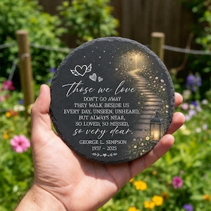 May include: A round black slate memorial plaque featuring a printed image of a stone path leading to a glowing light, a lantern, and flowers. The text reads: Those we love don't go away, they walk beside us every day, George L. Simpson 1937-2025.