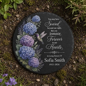 May include: A round black slate memorial stone featuring a printed illustration of blue and purple hydrangea flowers and a hummingbird. The text reads: You may have Soared beyond our sight, But you remain Forever in our Hearts, In Loving Memory Of Sofia Smith 1952-2026.