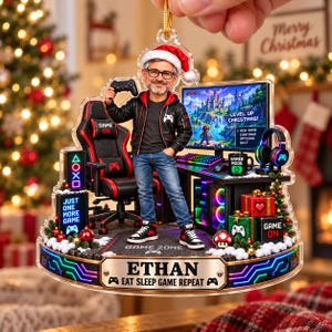 May include: A clear acrylic Christmas ornament featuring a man in a Santa hat standing in a gaming room. The scene includes a gaming chair, computer monitor with game text, a desk, headphones, and gift boxes. A plaque at the base reads ETHAN EAT SLEEP GAME REPEAT.